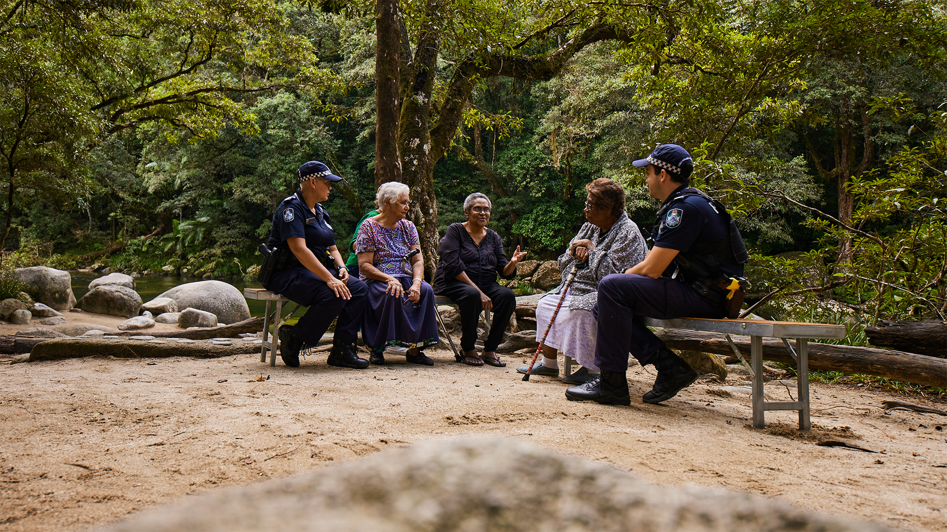 A group of First Nation's People's having a yarn with Queensland Police officers sat by a creek in the forest.